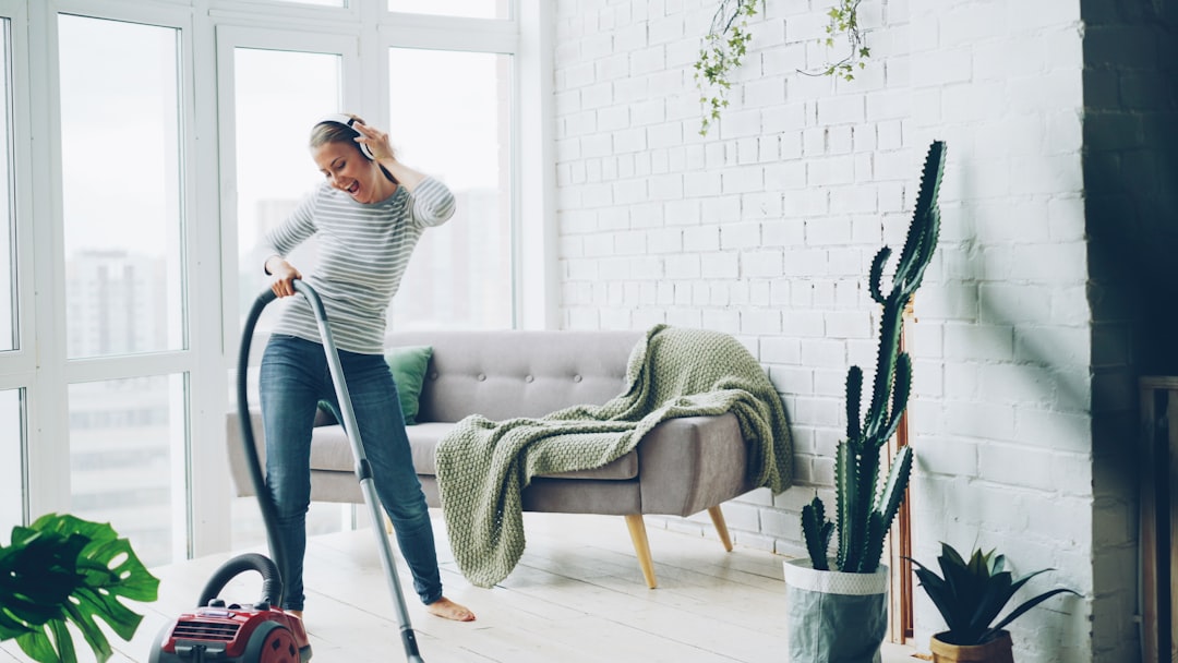 Cheerful blond housewife is using vacuum cleaner during routine clean-up at home, listening to music through headphones, singing and dancing. Technology and young people concept.