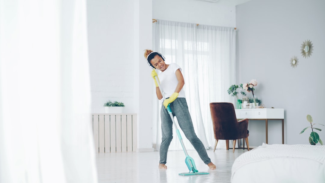 Happy housewife is doing housework and listening to music wearing modern wireless headphones, pretty young lady is singing in mop and dancing having fun at home.